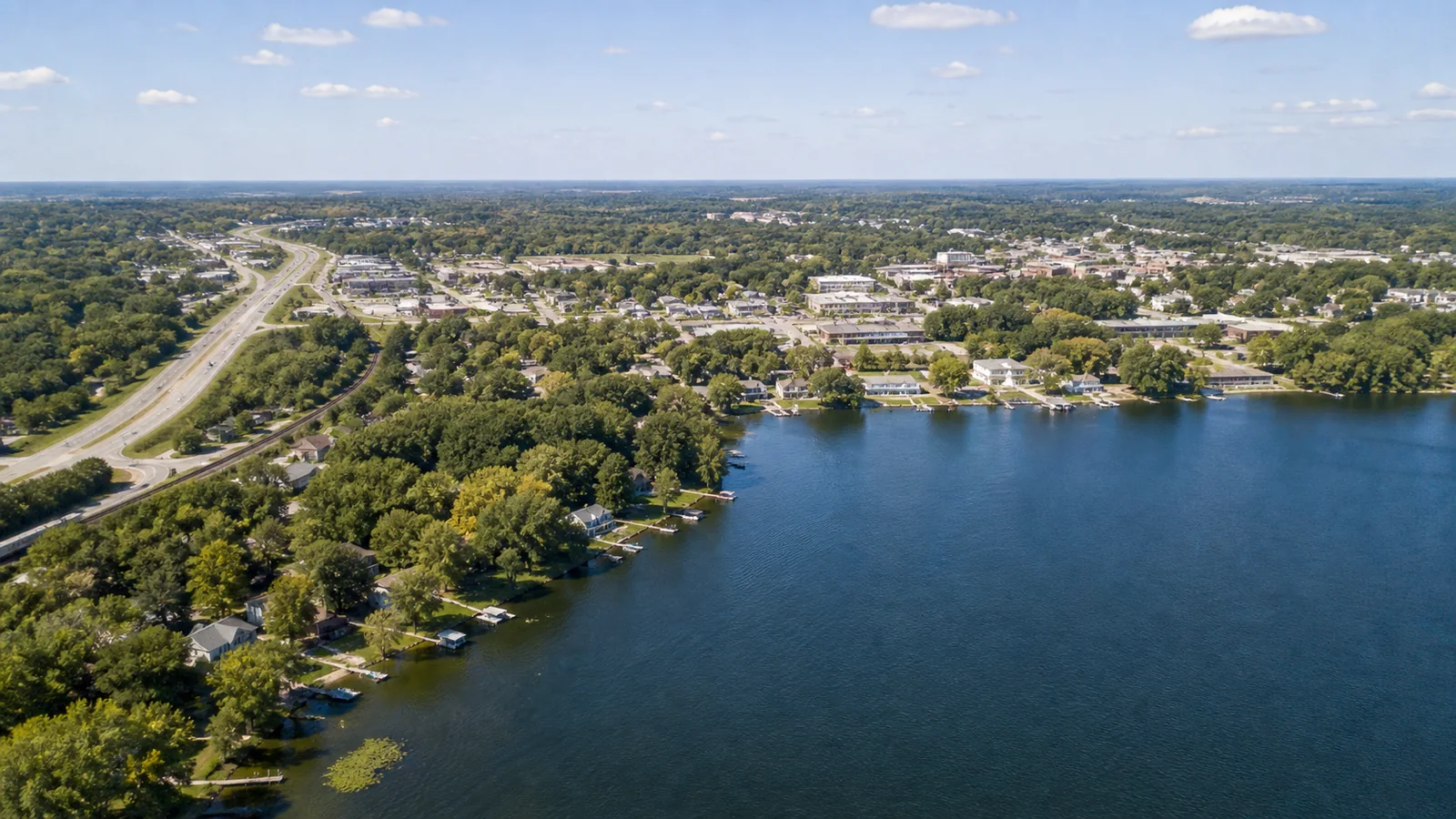 Editorial wide view of Forest Lake shoreline, public access, wooded neighborhoods, compact downtown-scale blocks, highway connection, and employment edges in Forest Lake