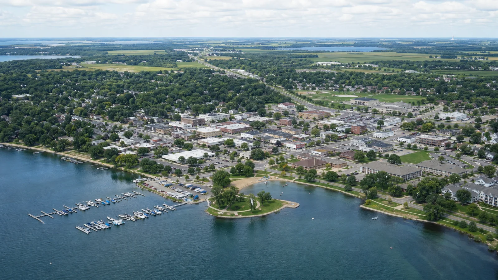 Editorial wide view of Lake Waconia shoreline and marina, public park, compact downtown, healthcare and business sites, neighborhoods, and agricultural edges in Waconia
